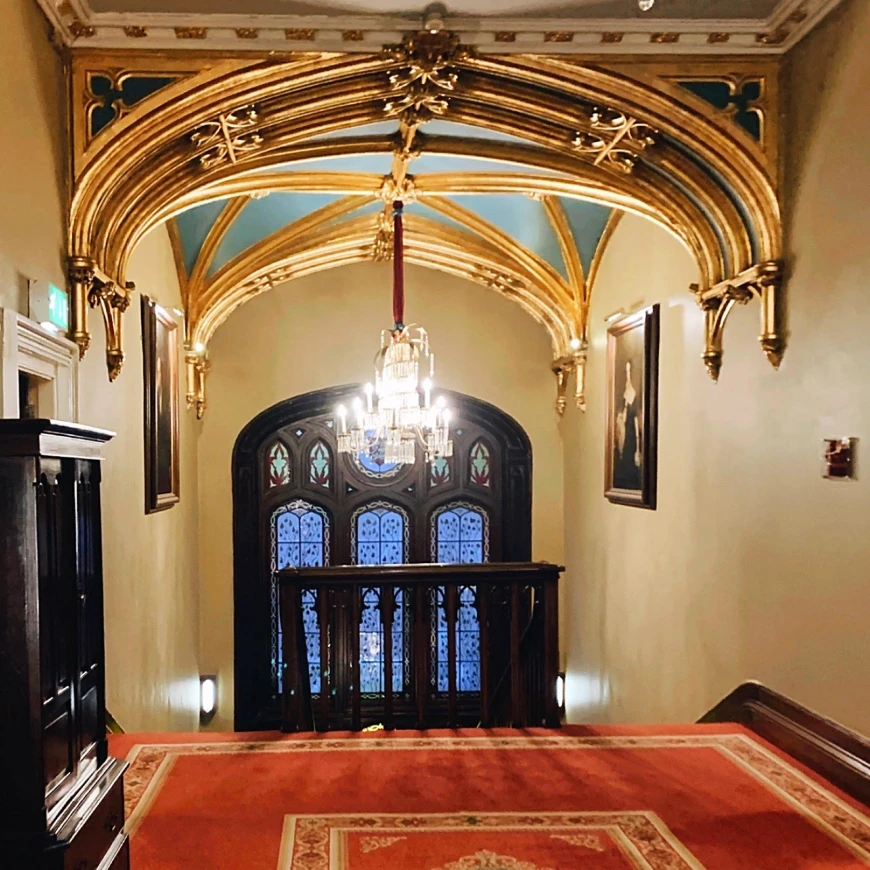 A view of a hallway with detailed gold paneling on the ceiling, a chandelier and stain glassed windows with wood trim. 