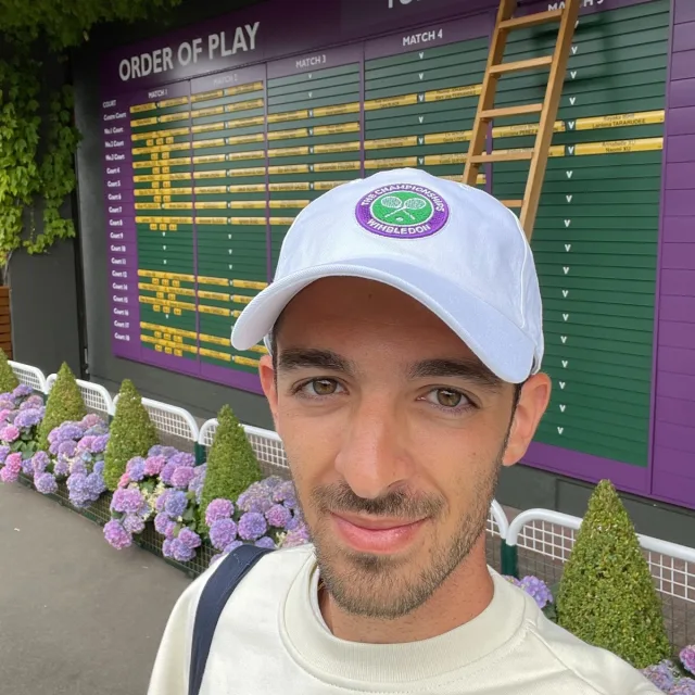 Travel Advisor Morris Hanono in a white Wimbledon hat with a purple and yellow scoreboard behind him and purple/green flowers.