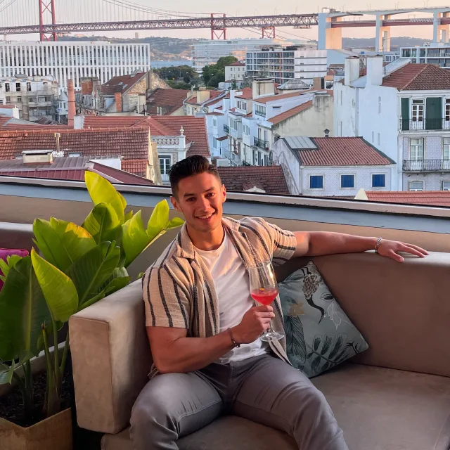 A man sitting in a lounge chair while holding a red drink in front of a view of red roofed and white buildings. 