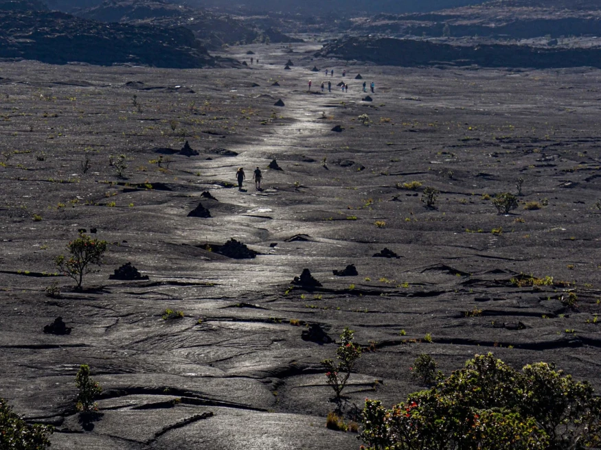 people walk on the black rock surface of a volcano in the sun