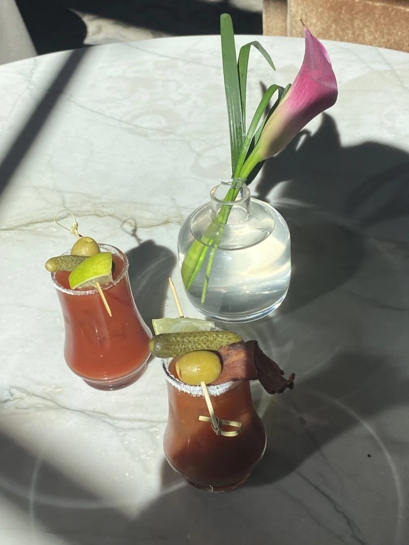 A white, round table set with two bloody marys and a pink flower in a vase.