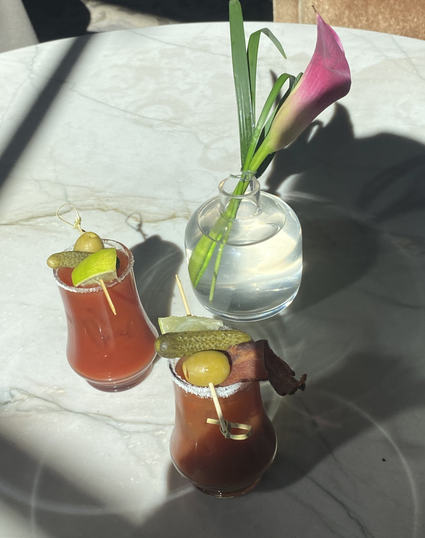 A white, round table set with two bloody marys and a pink flower in a vase.