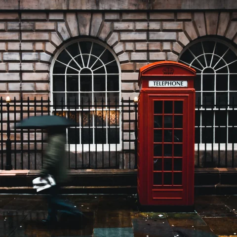 woman with a black umbrella walking by a red telephone booth on a rainy day