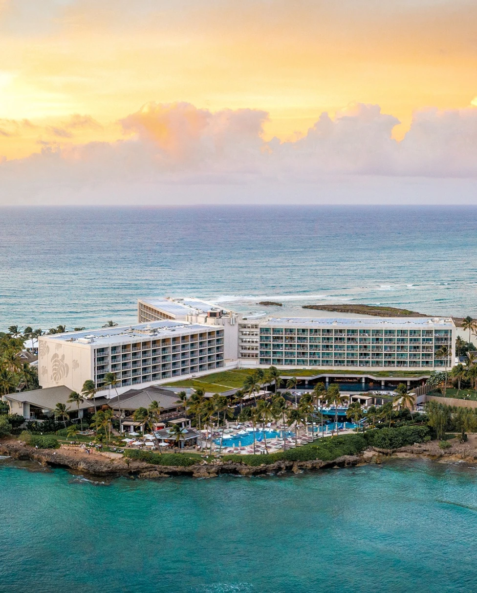 An aerial view of Turtle Bay Resort, a large white hotel with a bay in front and Oahu beaches beyond, at sunset.