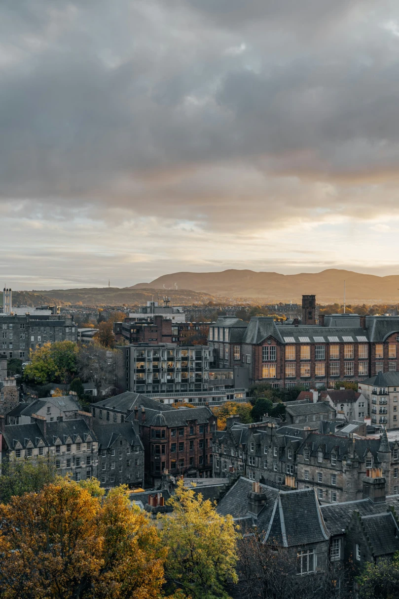A cityscape at sunset, featuring a blend of buildings and distant mountains under a cloudy sky, bathed in the soft evening light.