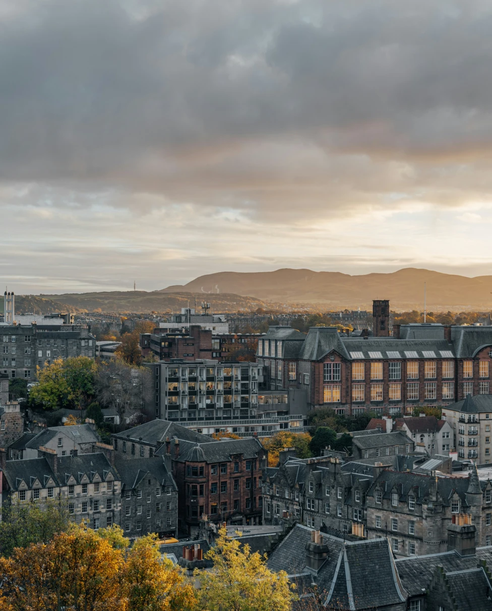 A cityscape at sunset, featuring a blend of buildings and distant mountains under a cloudy sky, bathed in the soft evening light.