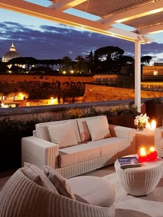 The rooftop patio at the Villa Agrippina Gran Melia, with white furniture overlooking trees and a Roman building at night. 