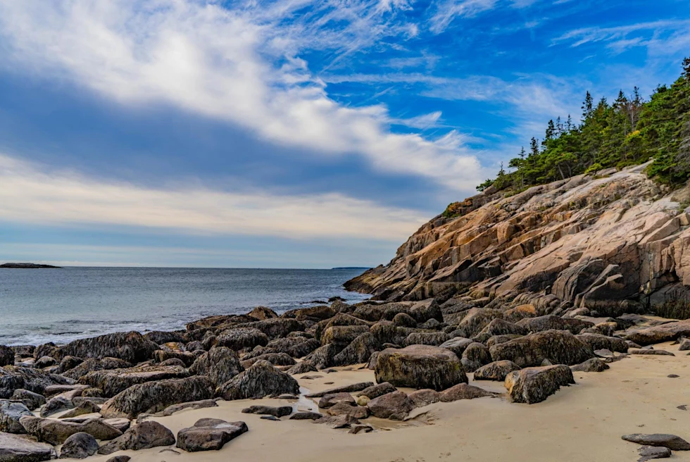 beach with large rocks dotting the shore and rising rocky cliffs to green forest on a sunny day