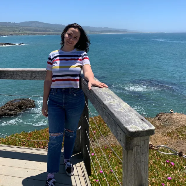 Travel Advisor Erica Simion Philips wearing white shirt with multi colored stripes and jeans with sea in the background. 
