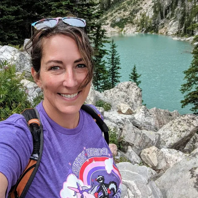 Travel Advisor Janet Louise in a purple shirt in front of a blue lake with rocks and trees.
