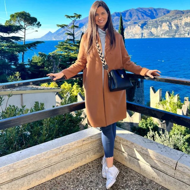 Picture of Lindsey wearing a camel colored coat with a black bag leaning against a black railing with blue water and mountains in the background