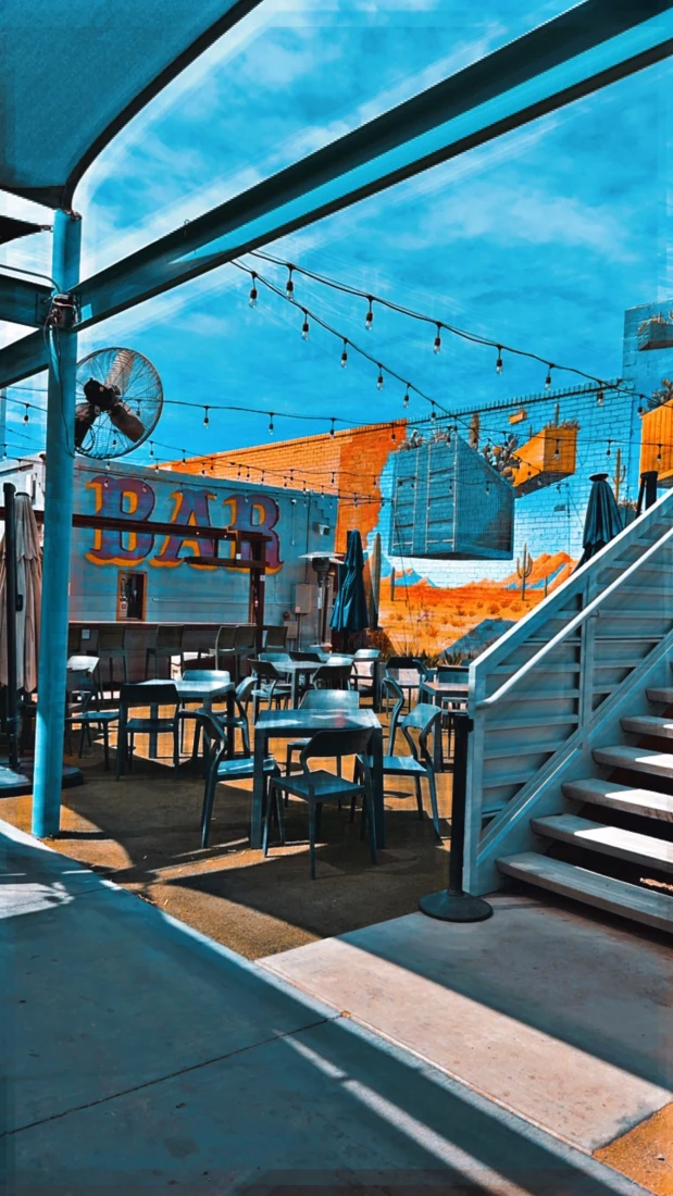 A view of an outdoor terrace with tables, chairs, a stairwell, string lights and a wall with the word "BAR" painted on it. 