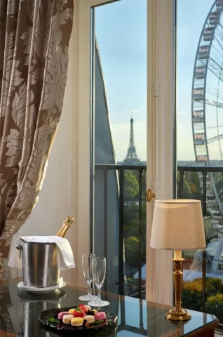 A view out the window of a room in the Regina Hotel Paris of the Eiffel Tower and Roue De Paris, with a table set with champagne and macarons in the foreground.