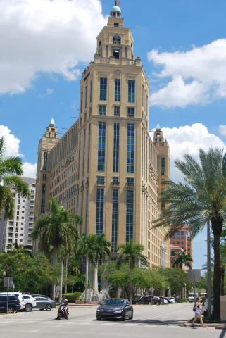 A picture of a tall beige color building surrounded by palm trees during daytime.