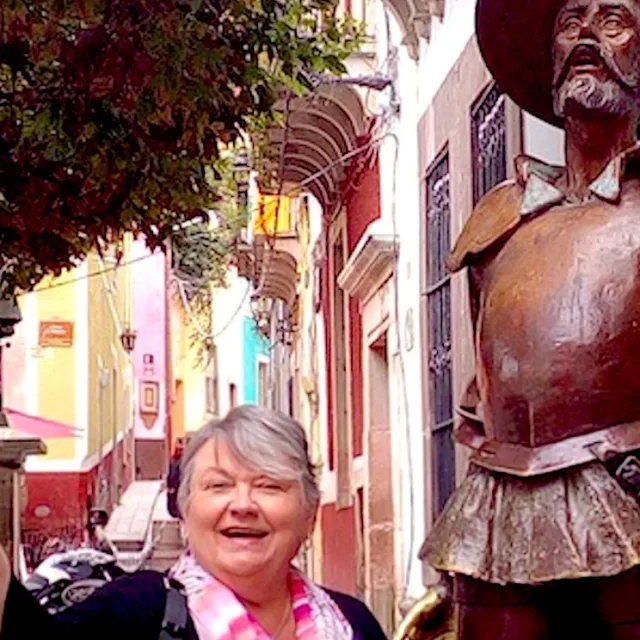 Travel Advisor Mary Manix stands next to a bronze statue on an old cit street with color buildings behind her 