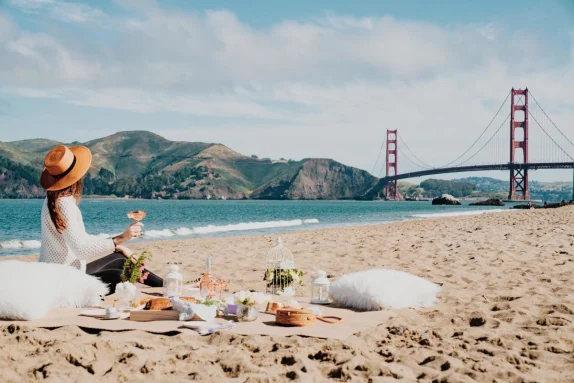 Woman wearing wide brimmed hat sits on the beach with a picnic overlooking the golden gate bridge in San Francisco
