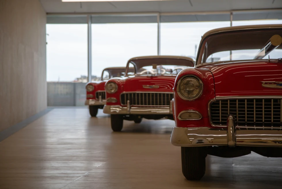 A row of three red cars in a light grey concrete room with windows at Fondazione Prada in Milan, Italy.