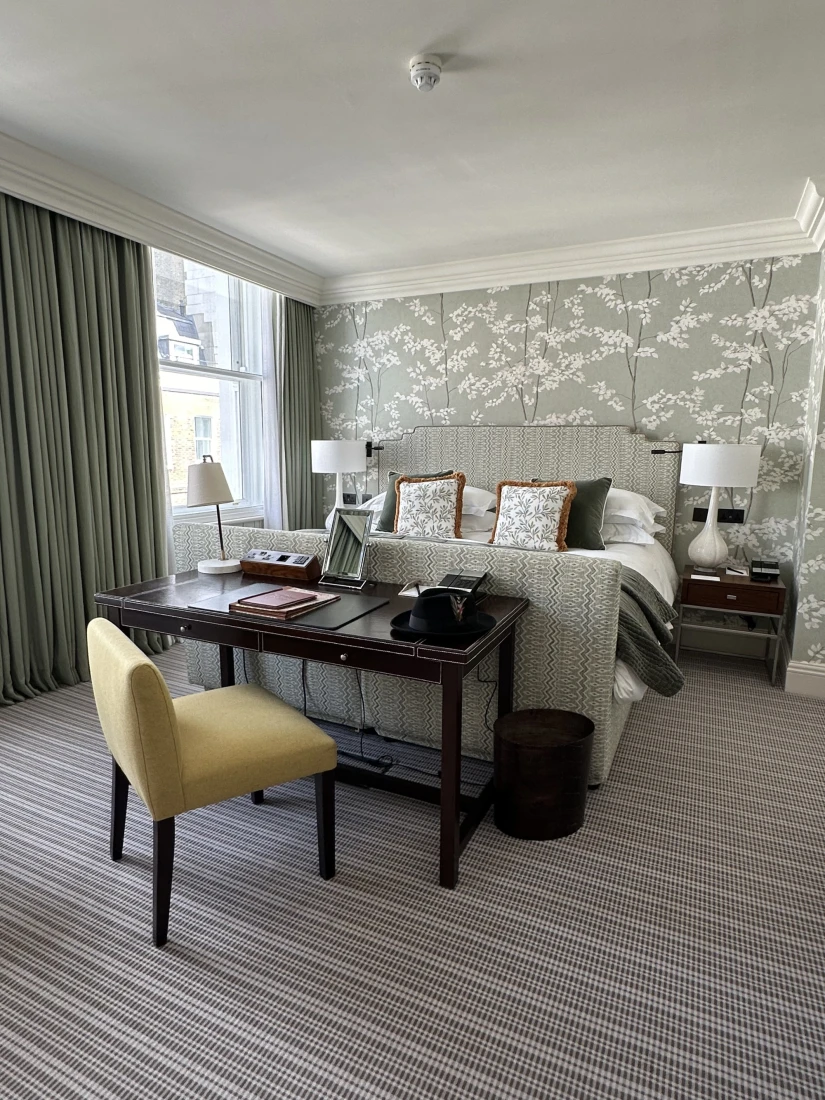 Junior Suite at Brown's Hotel showing a bed with gray and white patterned base and headboard, white bedside lamps, a desk with yellow chair and gray and white patterned wallpaper.