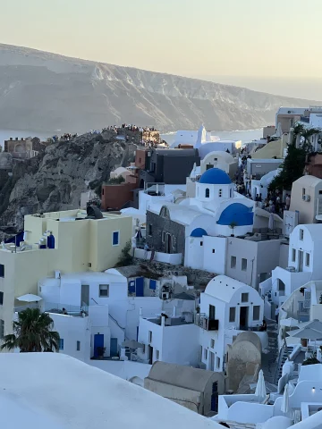 A picture of the beautiful white buildings of Greece with blue domed roofing nestled into a mountain, with the sea and another mountain view in the distance. 