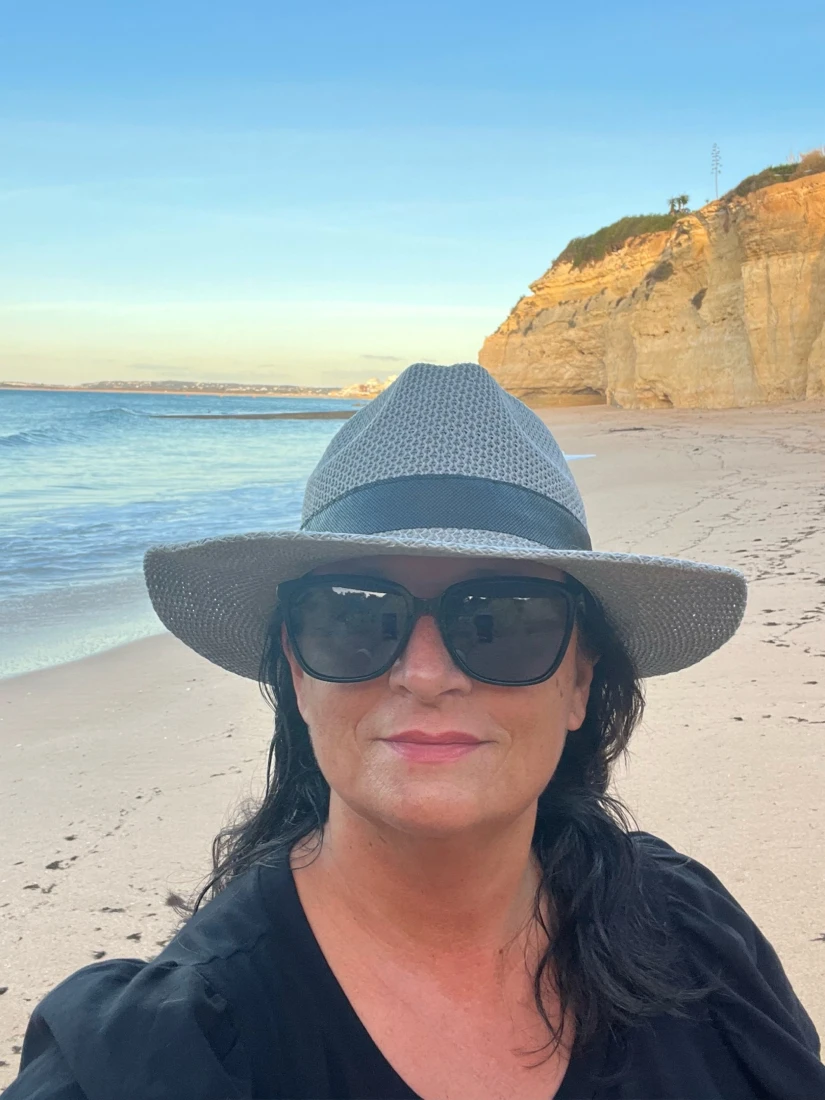 a woman in a hat and sunglasses smiles on a beach