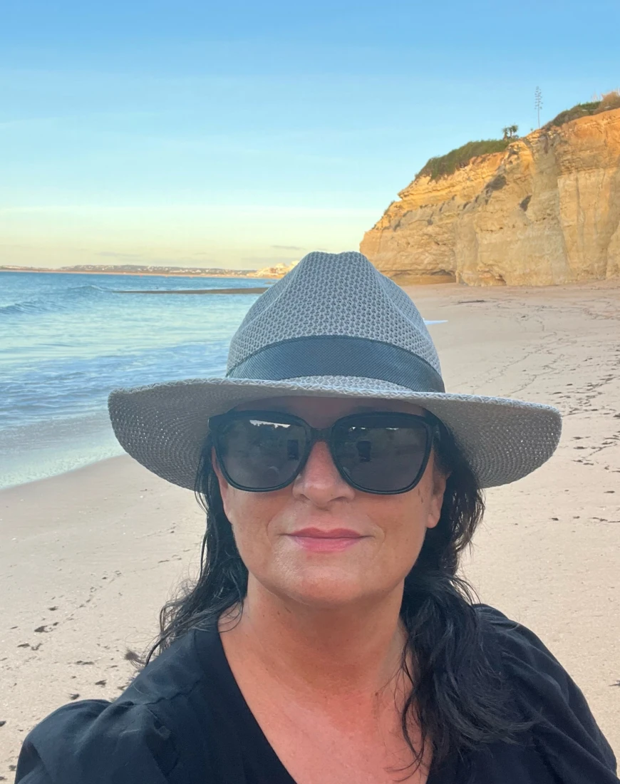 a woman in a hat and sunglasses smiles on a beach