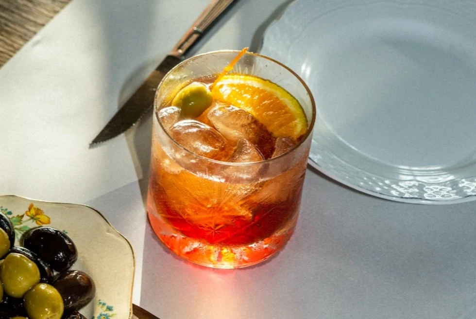small bowl of green and black olives near a red cocktail garnished with an orange slice