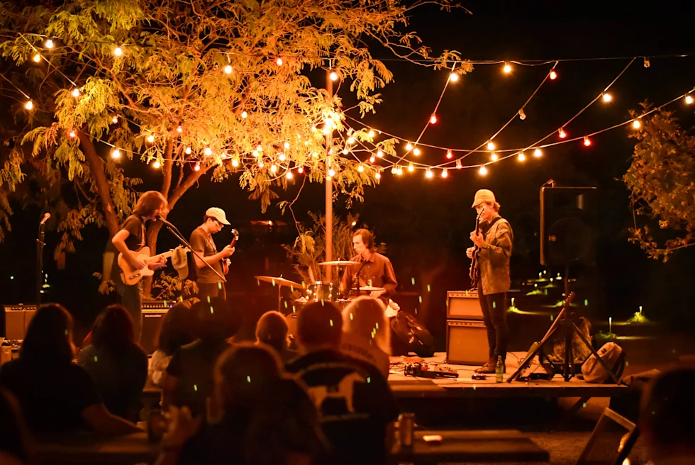 Four men in a band on stage with yellow string lights hanging from a tree playing guitars and drums for a crowd. 