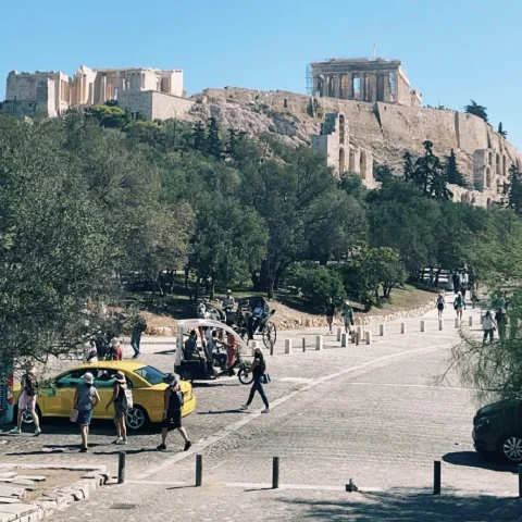 A street view of Athens with people, cars, trees and stone buildings in the distance. 