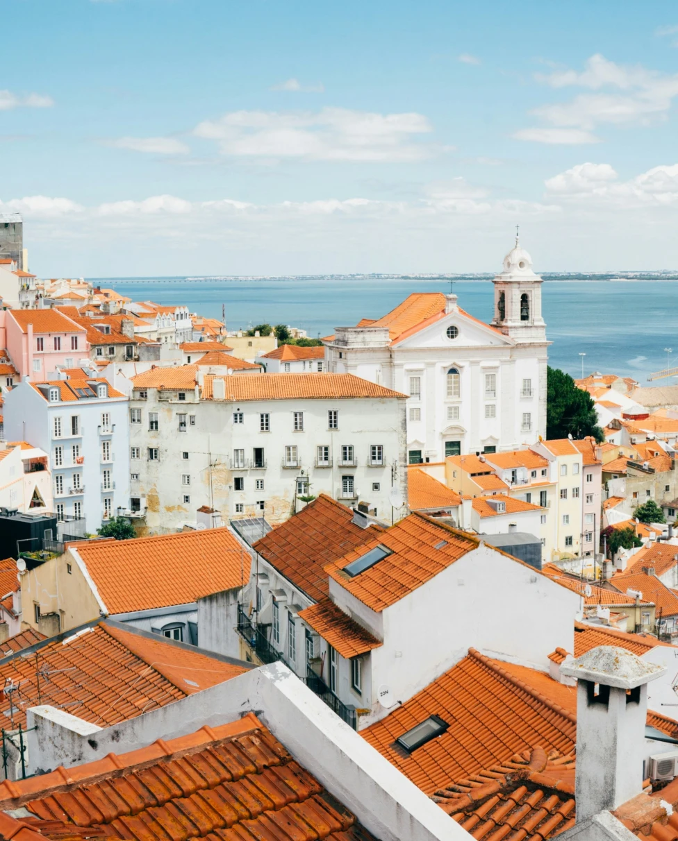 An aerial view of the landscape and architecture of orange roof houses in Lisbon with the ocean in the distance.