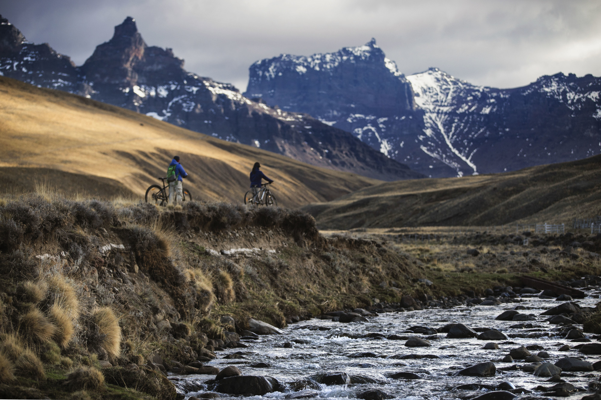 two bicycle riders on a path with a stream and mountains