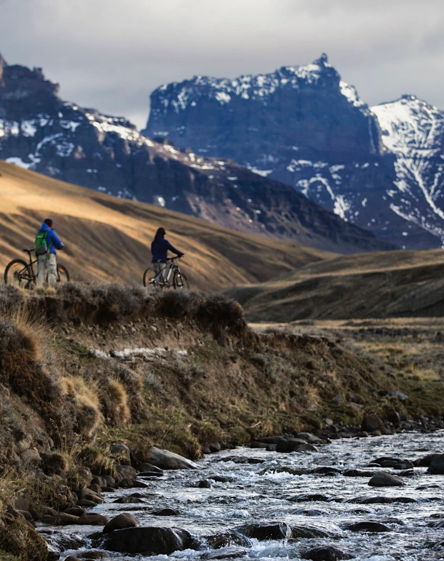 two bicycle riders on a path with a stream and mountains