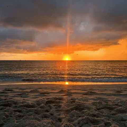 This is a serene and picturesque sunset at the beach. The sky is painted with hues of orange, yellow, and a touch of blue, indicating either dawn or dusk.