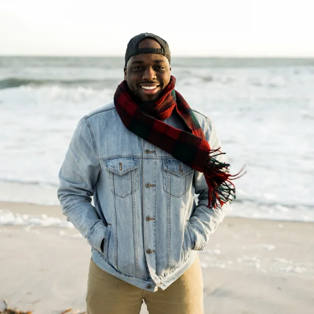 travel advisor Krystofer Henry in a light denim jacket and red scarf smiling on an ocean beach