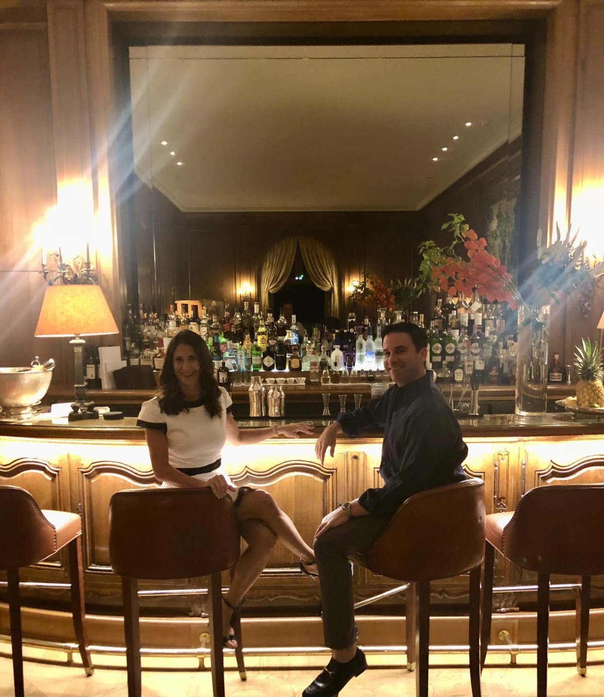 Chateau Bar LeRossini - a woman in a white cocktail dress and a man in a suit sitting at a wood-paneled bar with lamps