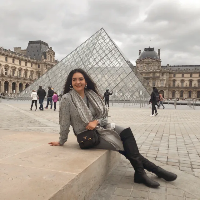 Travel Advisor Rosalva Gonzalez sits in front of the Louvre in Paris wearing a grey scarf, jumper and black boots