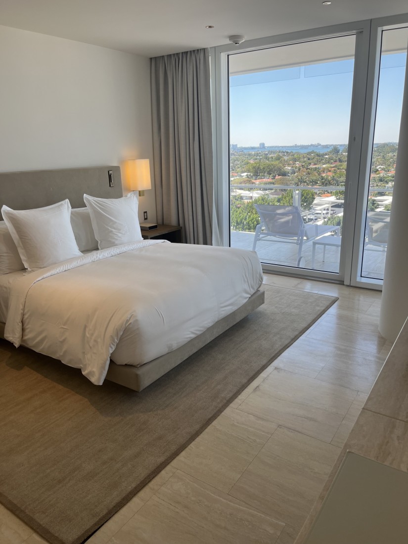 A hotel bedroom featuring a white bed with a beige headboard, a rug, wood flooring, nightstand with a lamp turned on and floor to ceiling windows looking out to a view of the city. 
