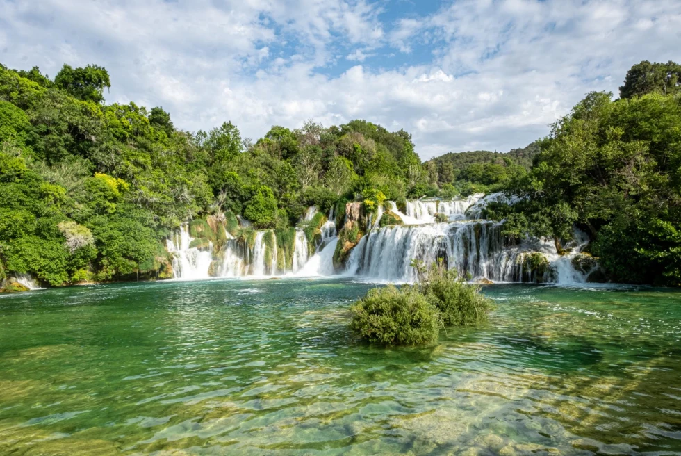 a natural waterfall pours into green shallow waters in a national park on a sunny day
