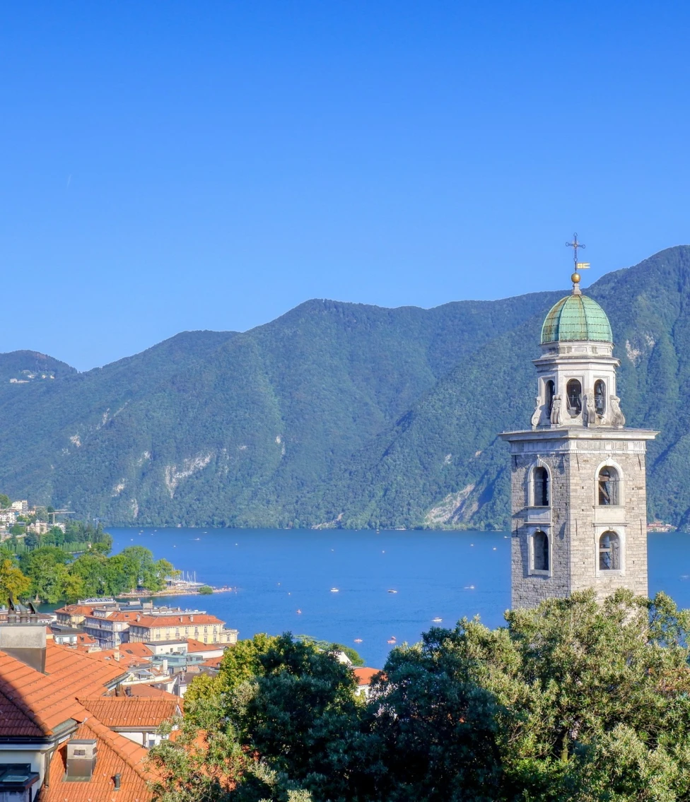 A view of Lake Lugano, a church steeple, and mountains during the daytime.