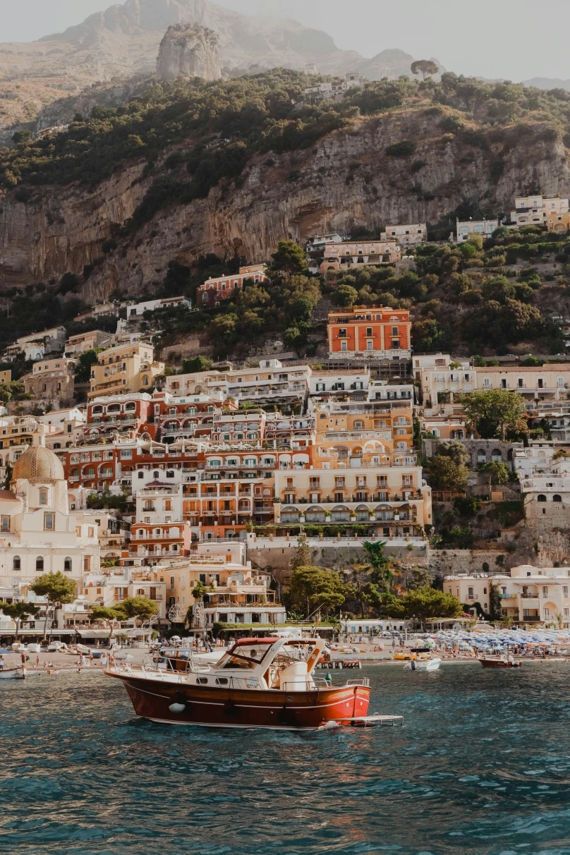 A view of a red and white boat sailing on the water in front of a coastal Italian town. 