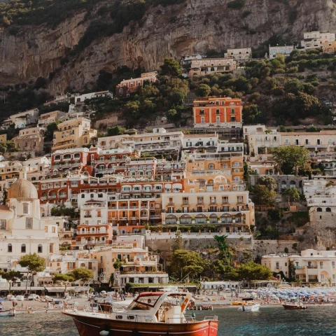 A view of a red and white boat sailing on the water in front of a coastal Italian town. 