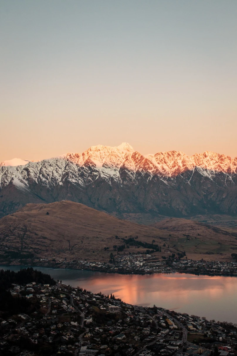 a town on a lake at the base of a snow topped mountain range at sunrise