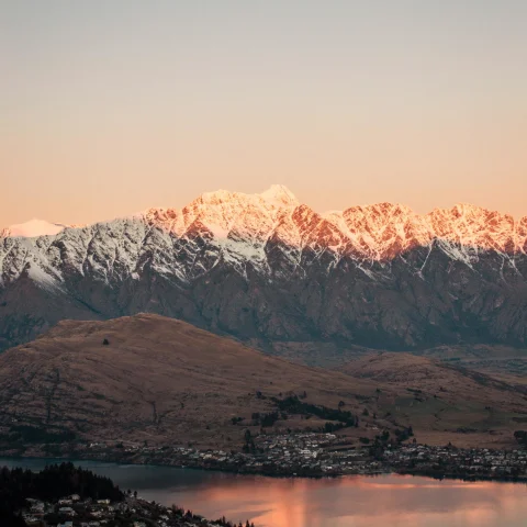 a town on a lake at the base of a snow topped mountain range at sunrise