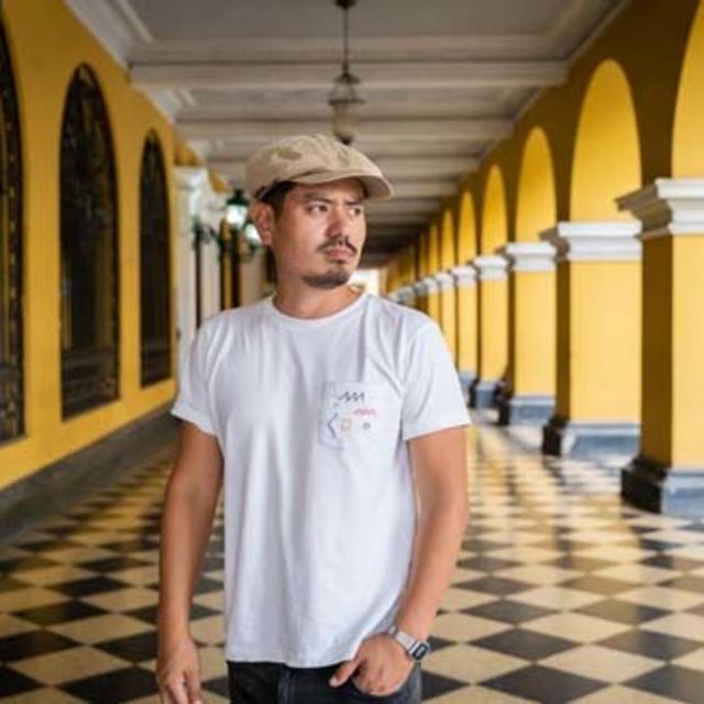 Fora travel agent Choji Itosu wearing tan hat and white shirt with yellow and black archways in the background