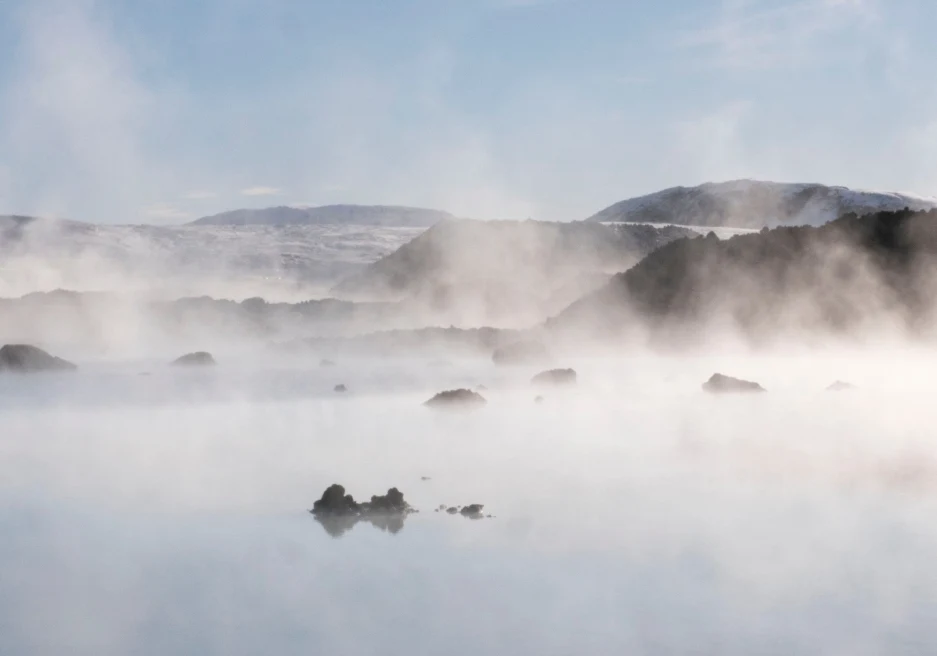 white steam rises from a natural pool with rocks peaking out and mountains in the distance