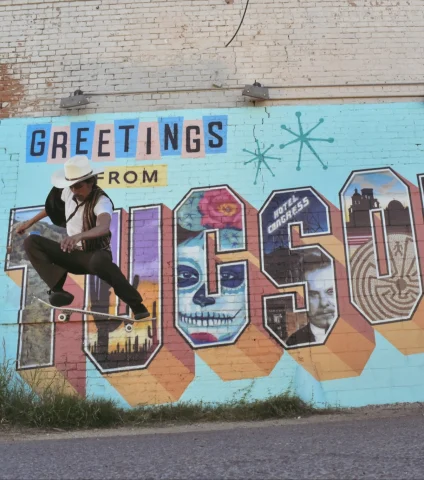 Man in a cowby hat on a skateboard midair, in front of a mural saying Greetings from Tucson.