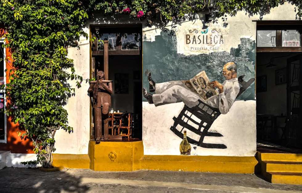 front of a pizzeria on a sunny day with green plants crawling up the walls and a mural with a man in rocking chair