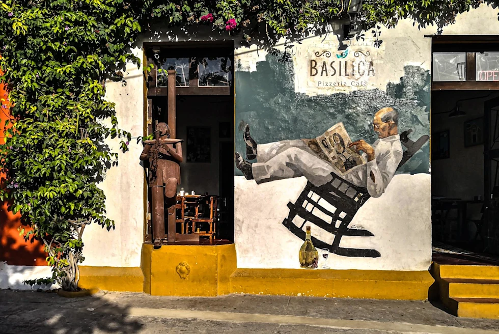 front of a pizzeria on a sunny day with green plants crawling up the walls and a mural with a man in rocking chair
