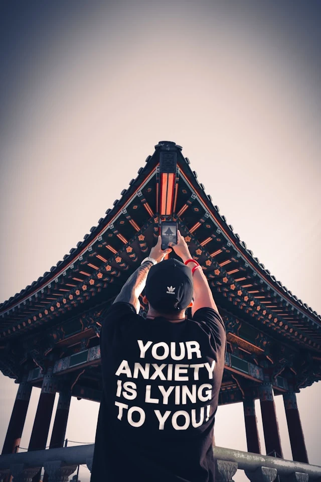 The back of a person taking a photo of a pagoda, wearing a shirt that says "Your anxiety is lying to you"