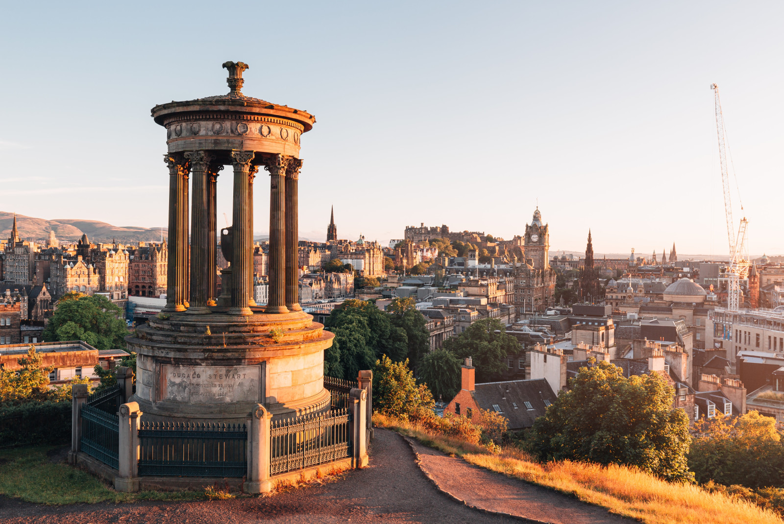 Travel Scotland to see old town Edinburgh overlooking the city with numerous ancient brown buildings, a tall white crane, and a brown, pillared monument at sunset. 