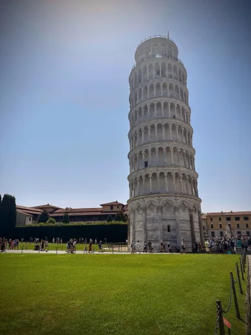 A view of the leaning tower of Pisa during the day time. There is a manicured, green lawn in the front and a clear blue sky in the background. 